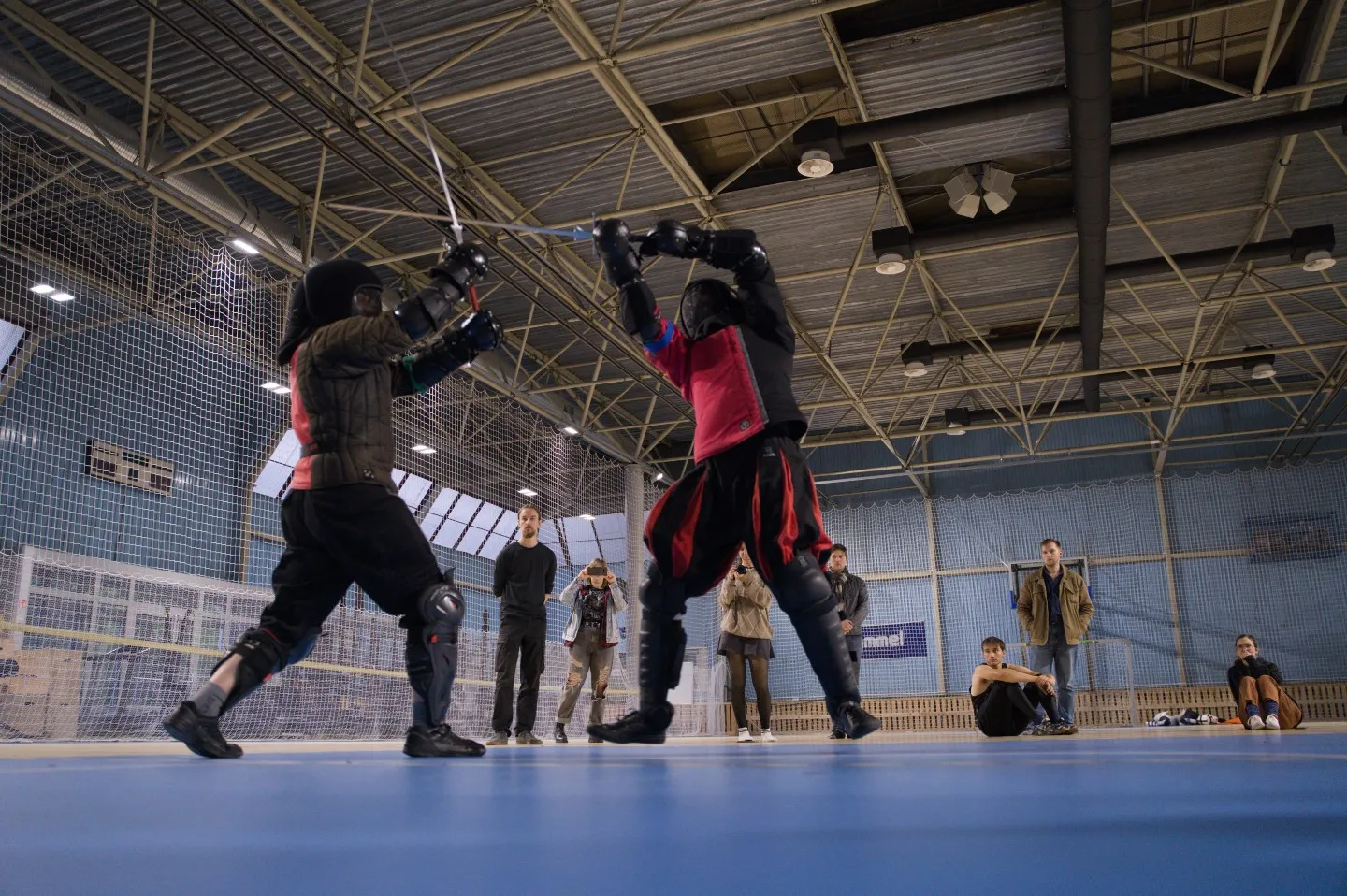 Two fencers in protective gear sparring with longswords in a sunlit hall