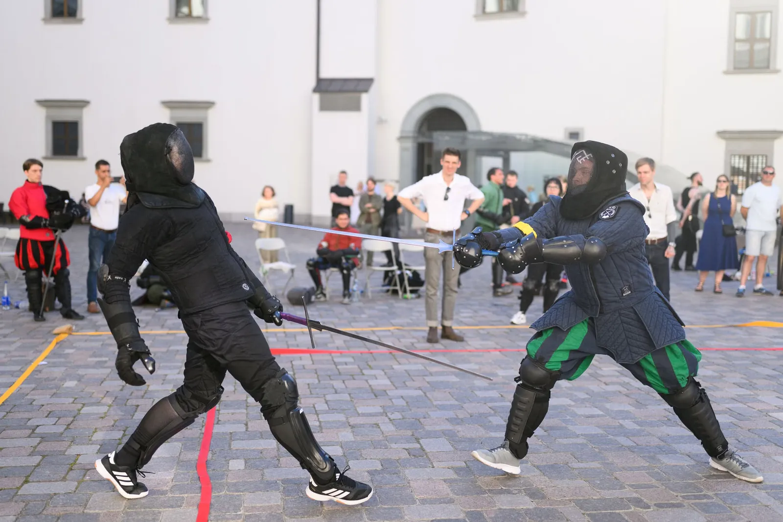 Longsword bout on a cobbled square by a historic building, watched by spectators
