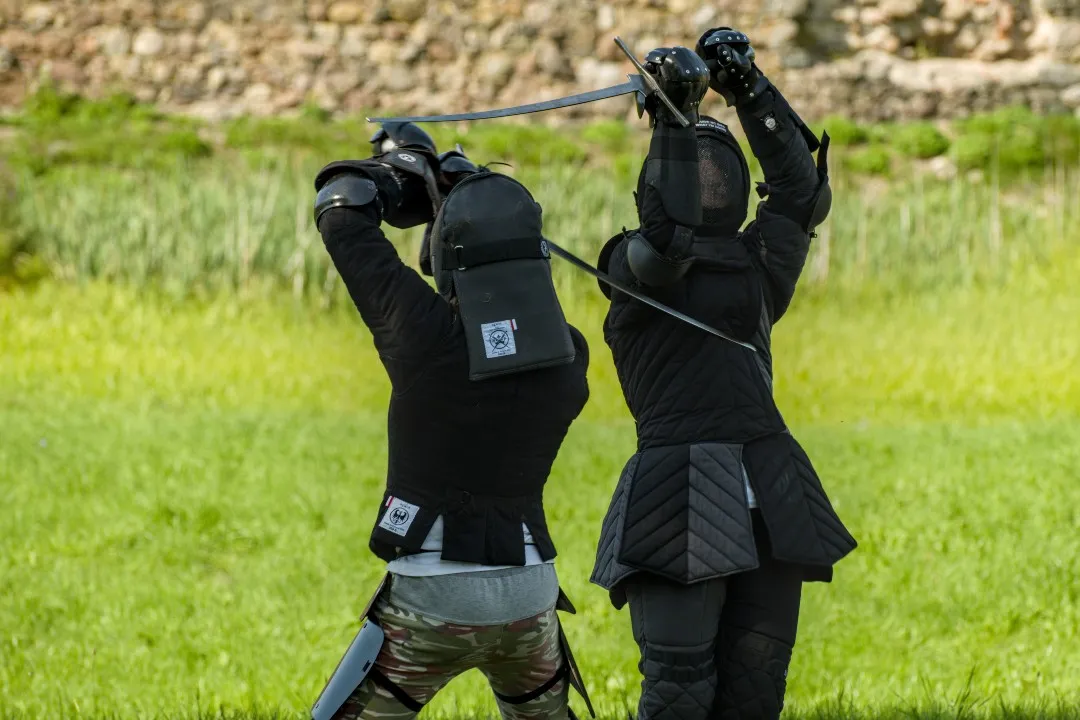 Two fencers crossing longswords in a meadow by an old stone wall